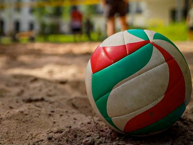 Nahaufnahme: Volleyball im Sand - dahinter das Netz und die Spieler/innen
