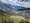 Panoramablick auf ein grünes Tal mit einem türkisfarbenen See und einer Siedlung, umgeben von bewaldeten Hängen und hohen Berggipfeln unter einem blauen Himmel mit Wolken. Ein Baumast im Vordergrund rahmt die rechte Bildseite ein.