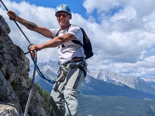 Ein lächelnder Mann mit Helm und Klettergurt hält sich an einem Stahlseil fest, während er eine Felswand erklimmt. Im Hintergrund sind bewaldete Berghänge und Gipfel unter blauem Himmel zu sehen.