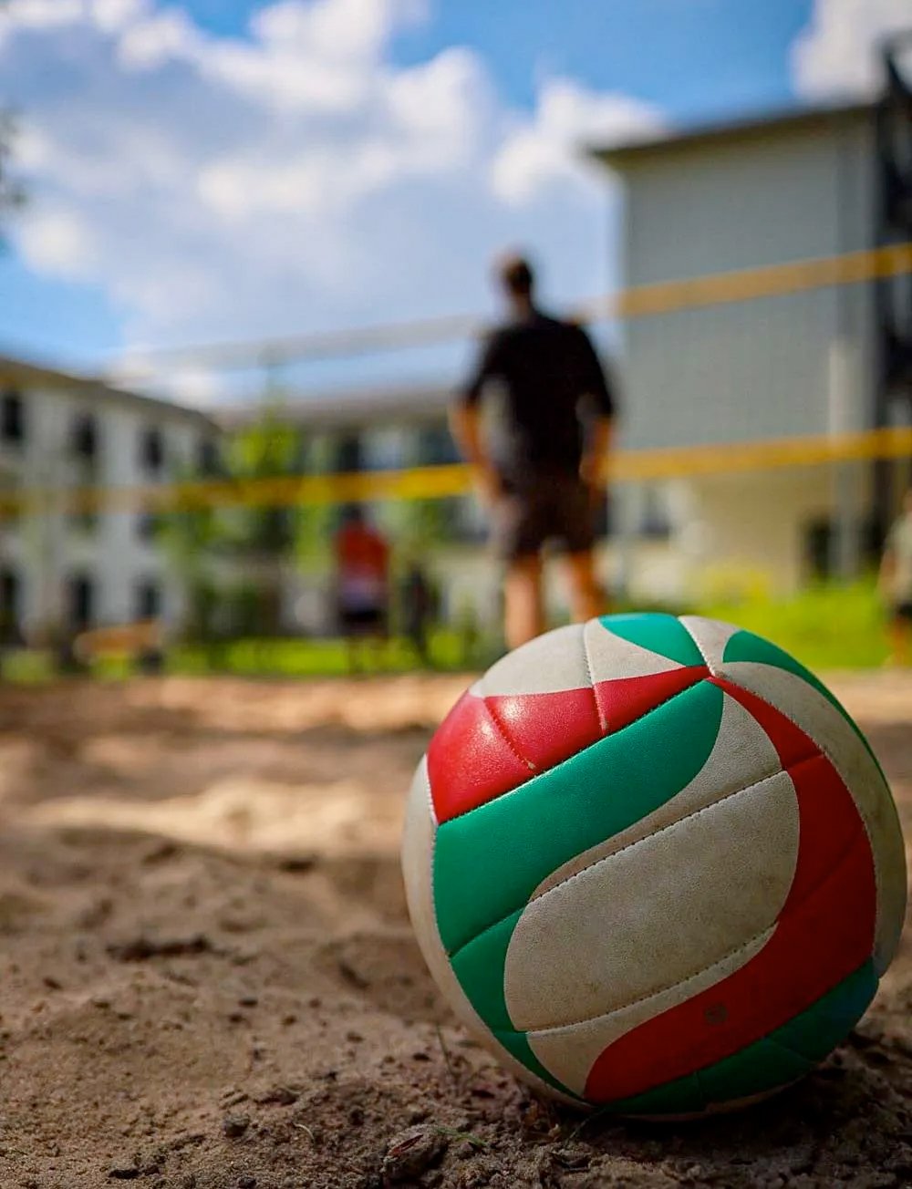 Nahaufnahme: Volleyball im Sand - dahinter das Netz und die Spieler/innen